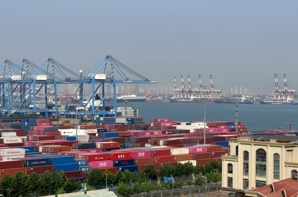 A sprawling container yard at a busy port with rows of colorful shipping containers, blue gantry cranes, and cargo ships docked in the background.