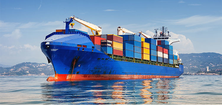 A large blue container ship loaded with colorful shipping containers sailing on calm waters, with a mountainous coastline in the background under a bright blue sky.