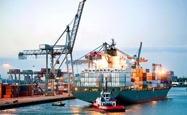 A large container ship docked at a port with gantry cranes, a tugboat assisting, and stacked shipping containers illuminated at dusk.