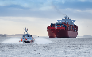 A small pilot boat speeding through choppy waters toward a large red container ship loaded with colorful cargo containers under a cloudy sky.