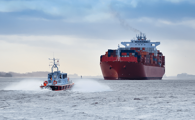 A small pilot boat speeding through choppy waters toward a large red container ship loaded with colorful cargo containers under a cloudy sky.