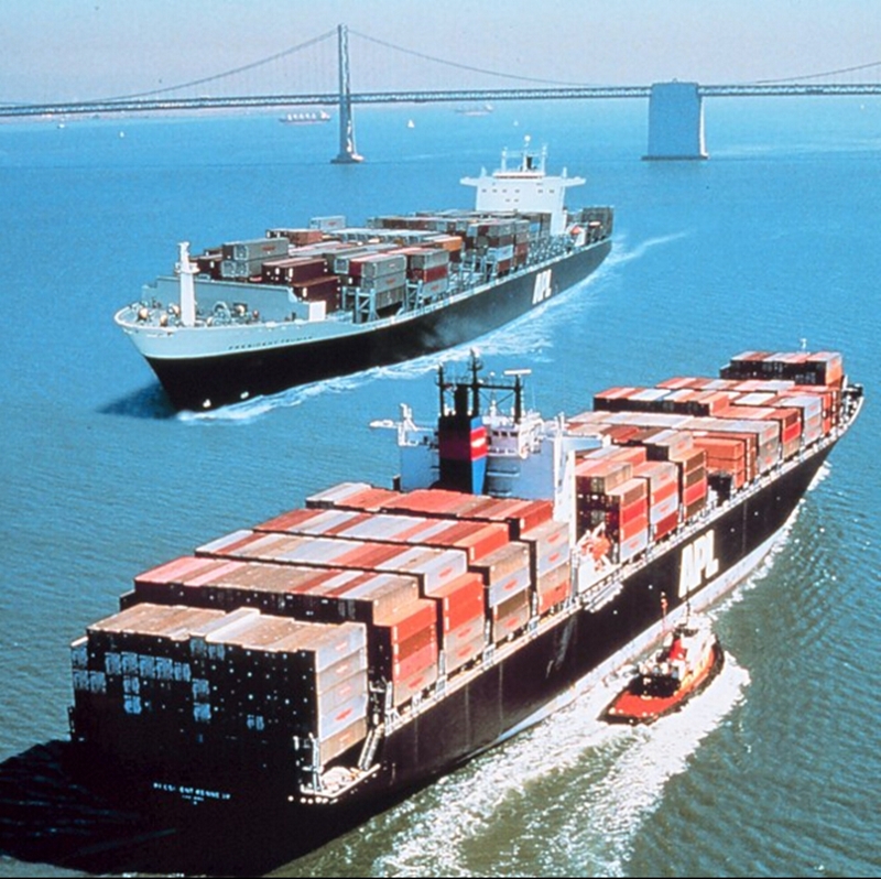 Two large container ships loaded with colorful shipping containers sailing in a bay, with a suspension bridge and a tugboat visible in the background.