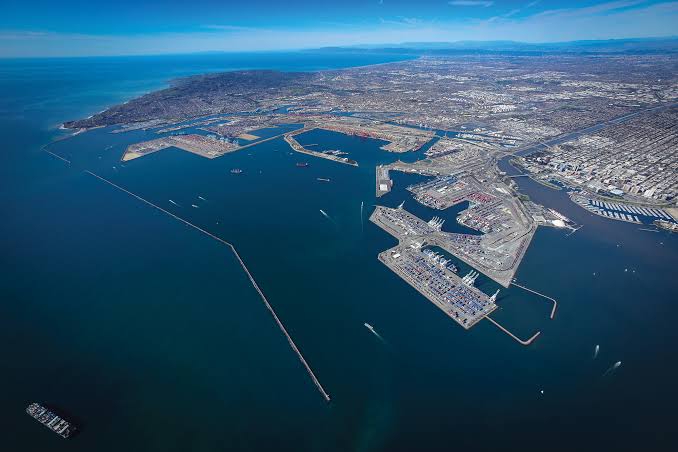 An aerial view of the Port of Long Beach in California, showing its massive harbor, breakwater, shipping terminals, and the surrounding city and coastline.