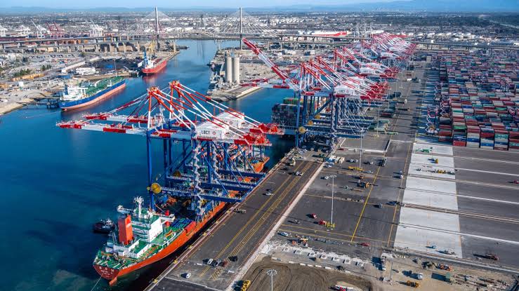 An aerial view of the Port of Los Angeles with a large container ship docked, massive gantry cranes, and extensive stacks of shipping containers.